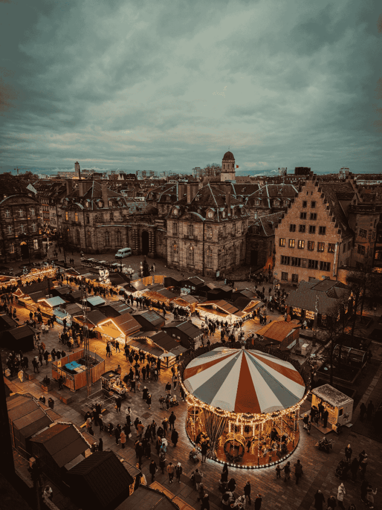 A bird’s-eye view of Strasbourg’s Christmas market shows glowing stalls, a carousel, and crowds gathering at dusk.
