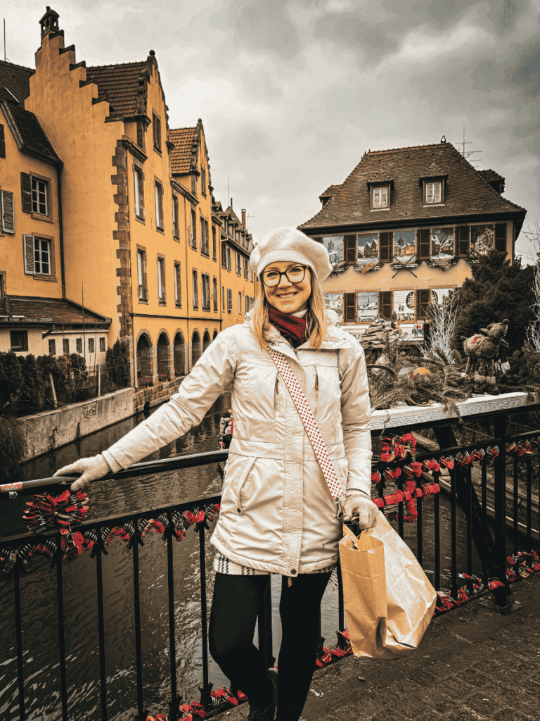 Kate smiles on a bridge in Colmar, with festive locks on the railing and colorful historic houses behind her.