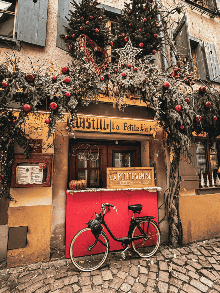 A bicycle leans against the festive storefront of La Petite Venise in Colmar, decorated with evergreens and ornaments.