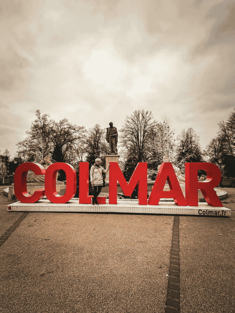 Kate stands beside the giant red Colmar sign in front of a statue (right).