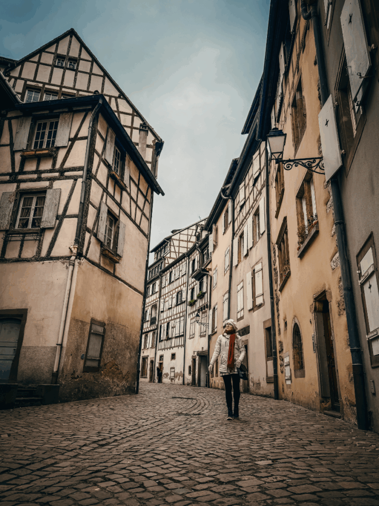 Kate walks alone down a quiet cobblestone street lined with old half-timbered buildings in Strasbourg.