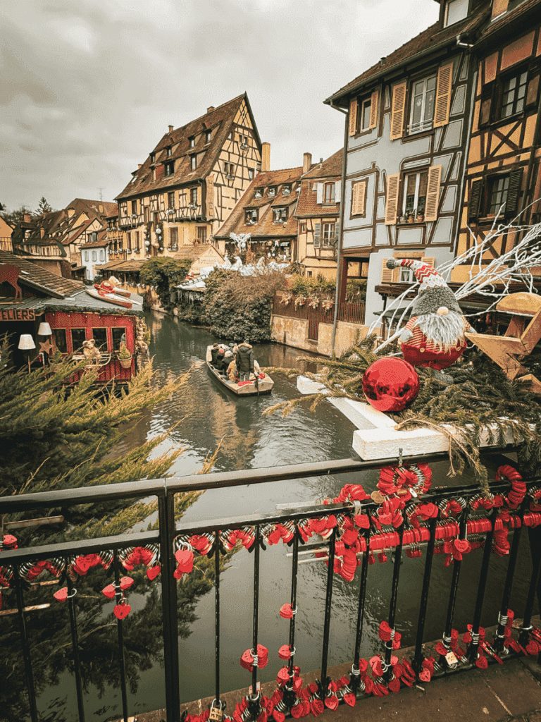 A festive canal scene in Colmar shows half-timbered houses, Christmas decorations, and a small boat gliding under a bridge lined with red love locks.
