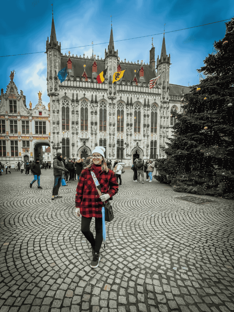 Kate stands in front of Bruges’ Gothic City Hall, smiling with festive decorations nearby.
