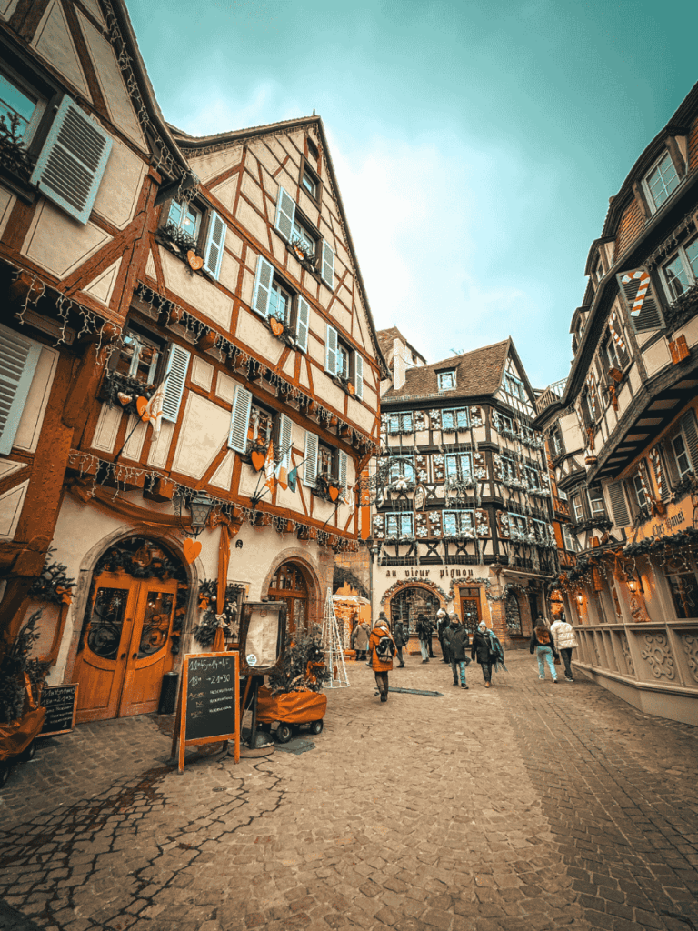 A charming street in Colmar is lined with half-timbered houses decorated for Christmas, with people strolling along the cobblestones.