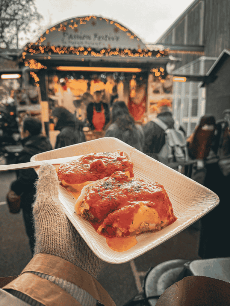A hand holds a plate of stuffed pasta with tomato sauce at a festive Strasbourg market stall.