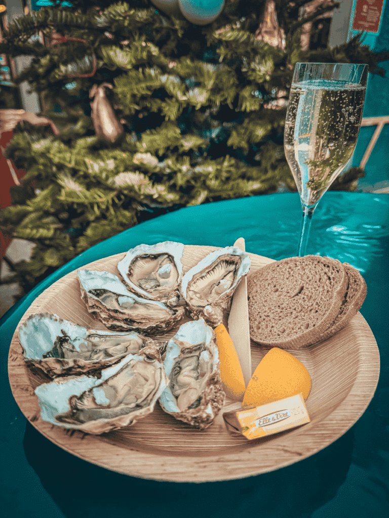 A plate of fresh oysters with lemon, bread, and butter is served with a glass of sparkling wine in front of a Christmas tree.