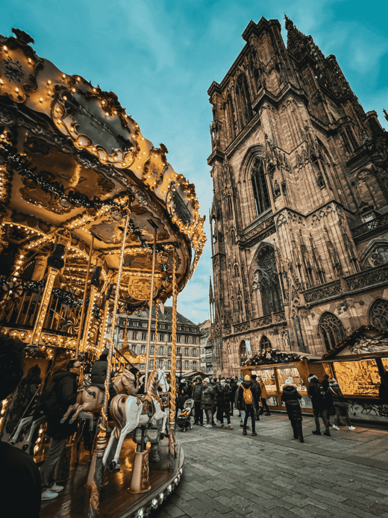 A brightly lit carousel with white horses spins in front of the towering Strasbourg Cathedral, surrounded by festive market stalls and visitors.