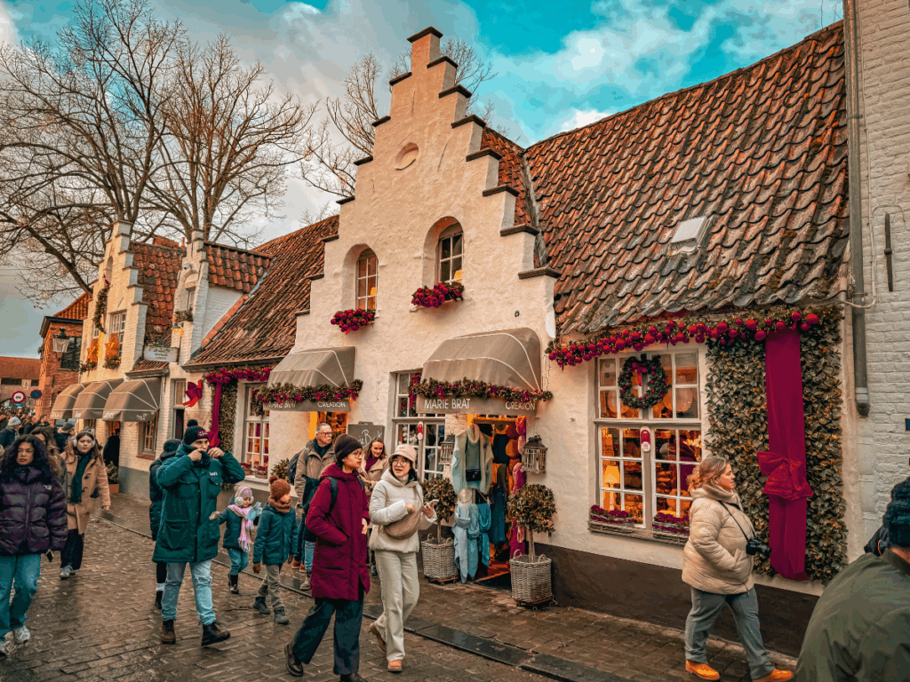 A historic brick building with red-trimmed windows stands along a canal in Bruges under a cloudy sky. You said: She Knows Alt Text said: People stroll past whitewashed shops decorated with wreaths and garlands in Bruges’ charming old town.