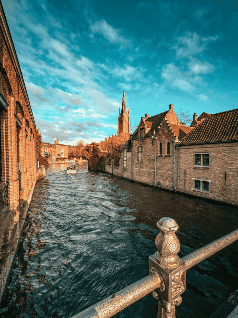 A canal in Bruges glistens in the sunlight with boats passing and the Church of Our Lady rising in the distance.