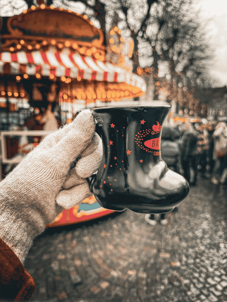 A gloved hand holds a black boot-shaped Bruges Christmas market mug in front of a carousel.