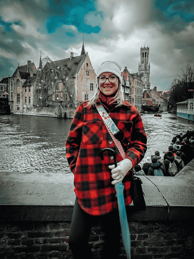 Kate stands with an umbrella by the water at Bruges’ Rozenhoedkaai, with historic buildings and the Belfry behind her.