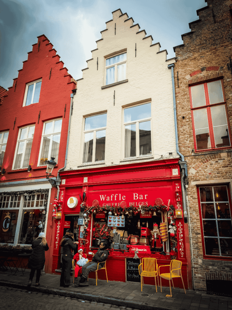 A bright red storefront called "Waffle Bar" in Bruges decorated for Christmas with people gathered outside.