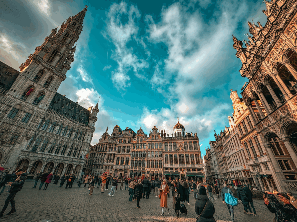 A historic brick building with red-trimmed windows stands along a canal in Bruges under a cloudy sky. You said: She Knows Alt Text said: People stroll past whitewashed shops decorated with wreaths and garlands in Bruges’ charming old town. You said: She Knows Alt Text said: Crowds gather in Brussels’ Grand Place, surrounded by ornate historic guildhalls under a bright blue sky.