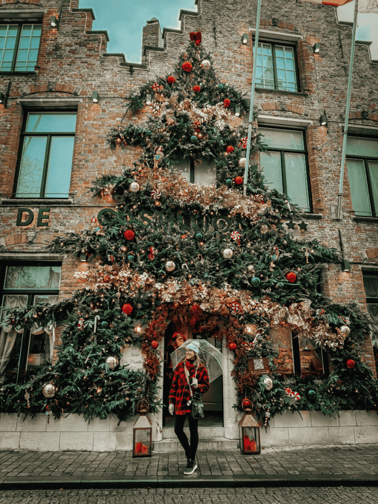 Kate stands with an umbrella in front of a Bruges building decorated with a giant Christmas tree display.