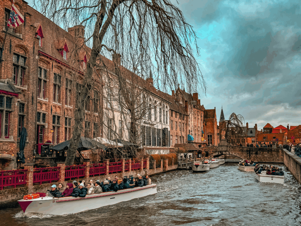 A historic brick building with red-trimmed windows stands along a canal in Bruges under a cloudy sky. You said: She Knows Alt Text said: People stroll past whitewashed shops decorated with wreaths and garlands in Bruges’ charming old town. You said: She Knows Alt Text said: Crowds gather in Brussels’ Grand Place, surrounded by ornate historic guildhalls under a bright blue sky. You said: She Knows Alt Text said: Tour boats filled with people glide along the canals of Bruges past historic brick buildings.