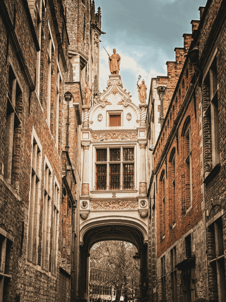 A golden-topped archway with ornate statues connects two historic brick buildings in Bruges.
