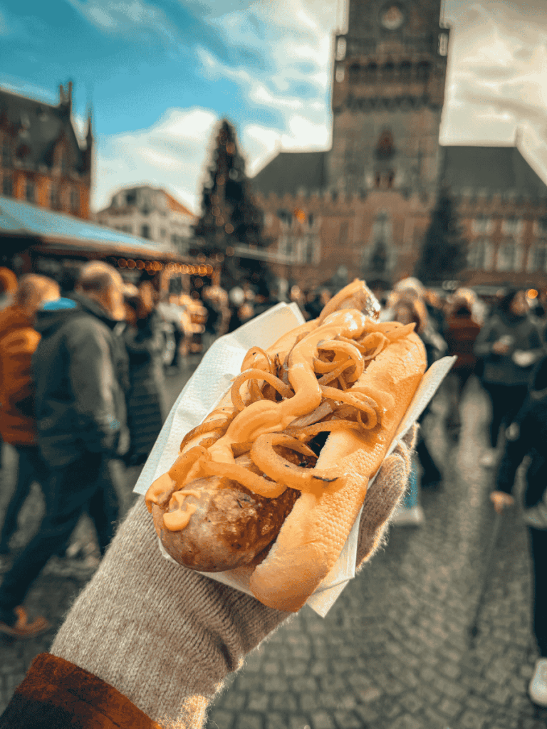 A sausage topped with mustard and onions is held up at a festive market in Bruges.