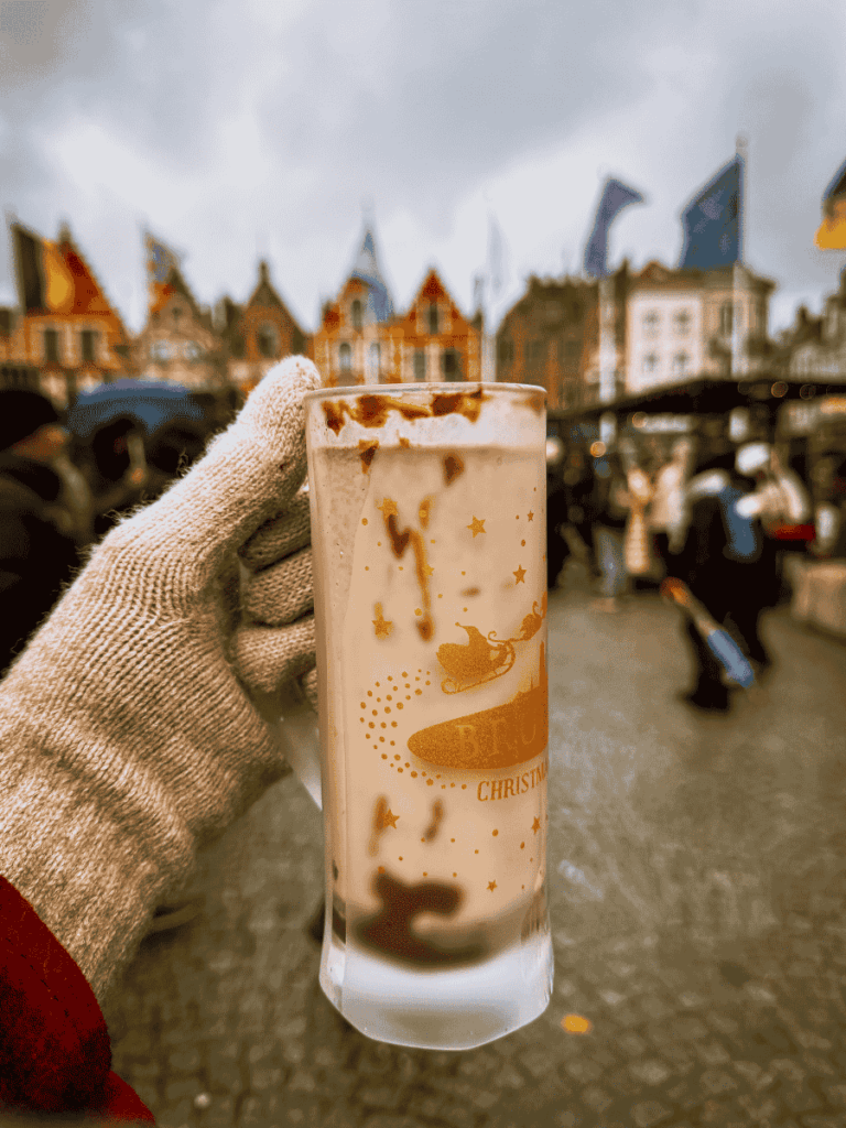 A gloved hand holds a Bruges Christmas Market mug filled with hot chocolate against a backdrop of colorful gabled houses.