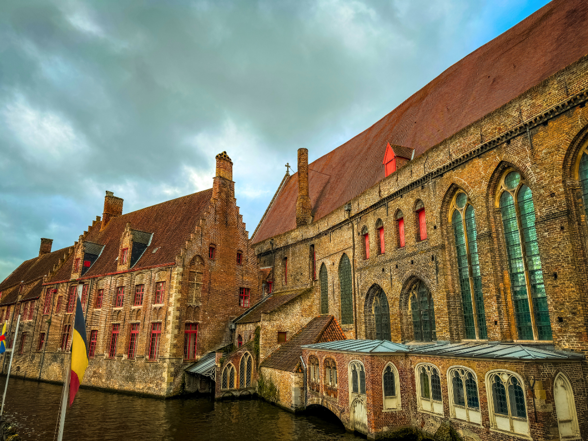 A historic brick building with red-trimmed windows stands along a canal in Bruges under a cloudy sky.