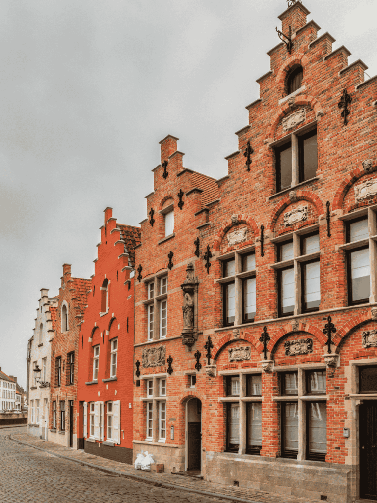Kate walks across Bruges’ main square toward a towering Christmas tree in front of the Gothic City Hall.

You said:
She Knows Alt Text said:

A row of traditional stepped-gable brick houses lines a quiet cobblestone street in Bruges.