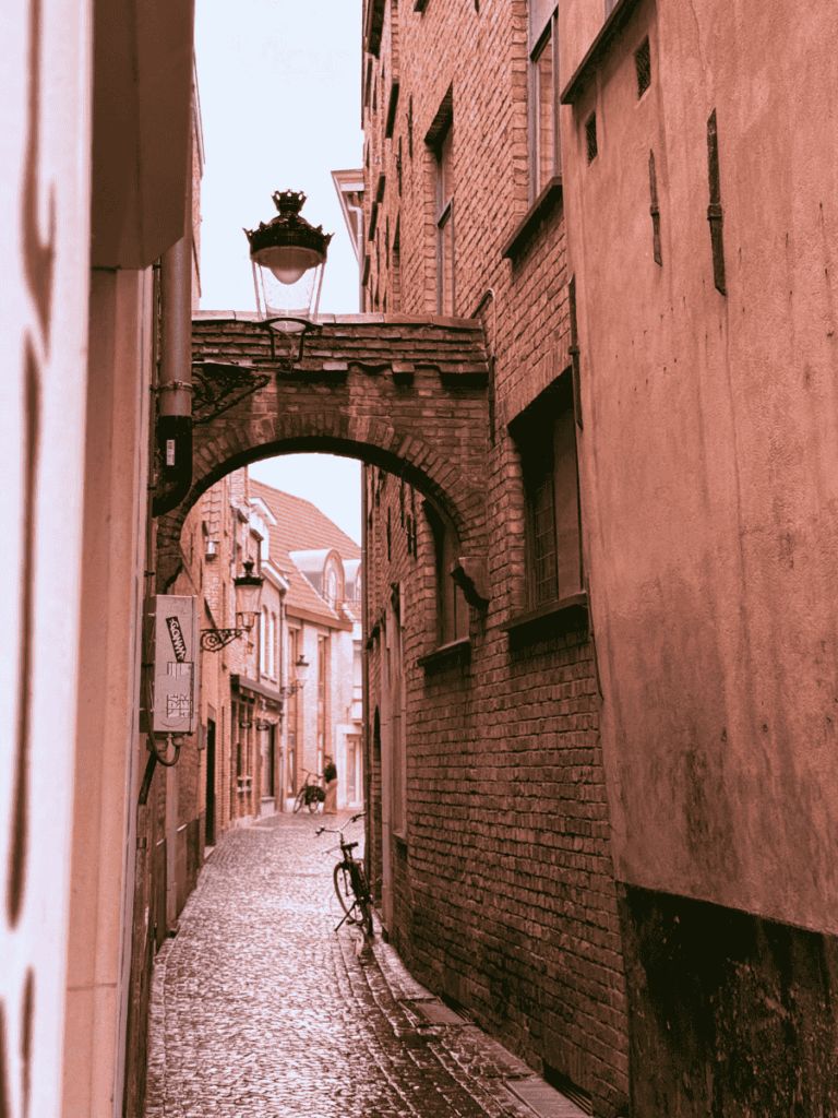 A narrow cobblestone alley in Bruges features brick walls, a hanging lantern, and a bicycle leaning against the side.