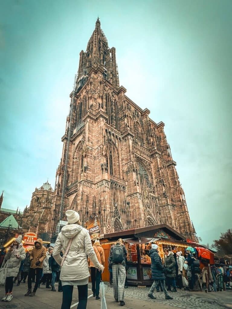 Kate looks up at the towering Strasbourg Cathedral, surrounded by Christmas market stalls and festive crowds.