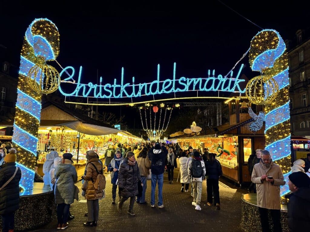 Crowds stroll under the glowing Christkindelsmärik sign at Strasbourg’s Christmas market, surrounded by festive lights and wooden stalls.