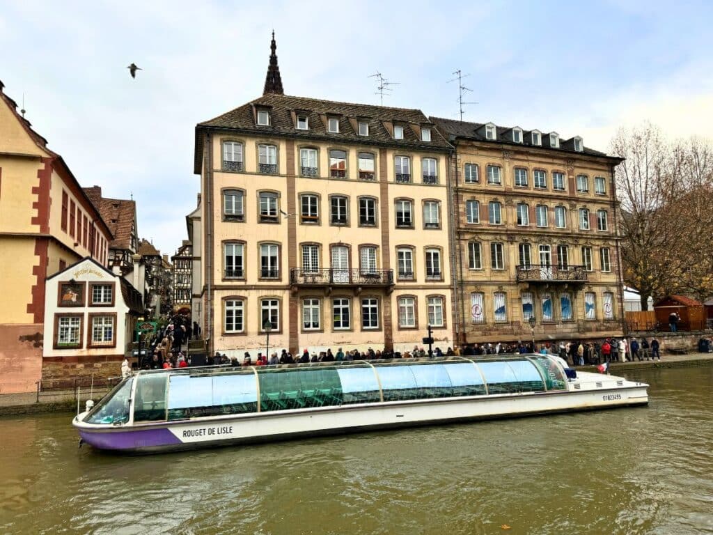 A glass-roofed sightseeing boat cruises along the river in Strasbourg, passing historic buildings and a crowd along the quay.
