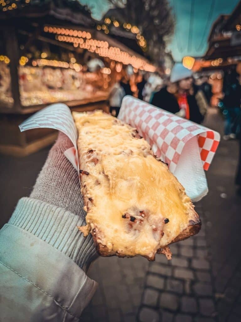 A hand holds up a slice of cheesy tartine topped with melted onions and lardons at a Christmas market stall in Strasbourg.
