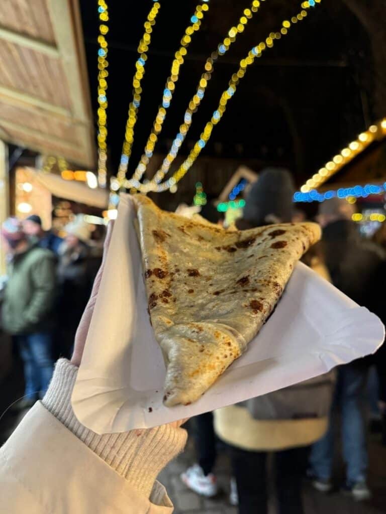 A warm folded crêpe is held up against a backdrop of twinkling lights and bustling Strasbourg Christmas market crowds.