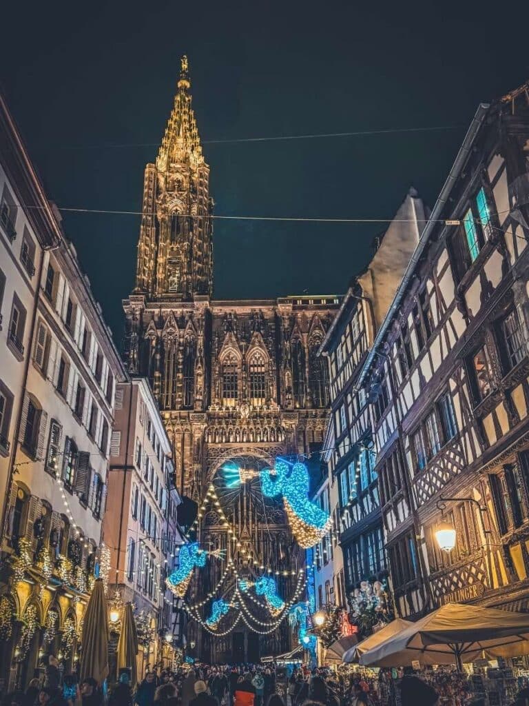 Strasbourg Cathedral shines brightly at night, framed by festive lights and half-timbered buildings on a lively street.
