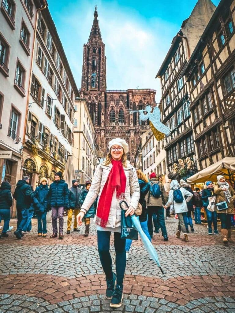 Kate smiles in front of Strasbourg Cathedral during the day, surrounded by crowds and holiday decorations.