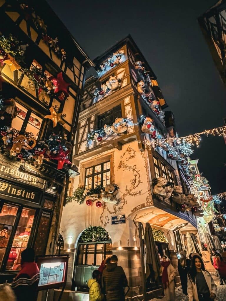 The same Strasbourg building glows at night, adorned with illuminated teddy bears, garlands, and festive holiday lights.