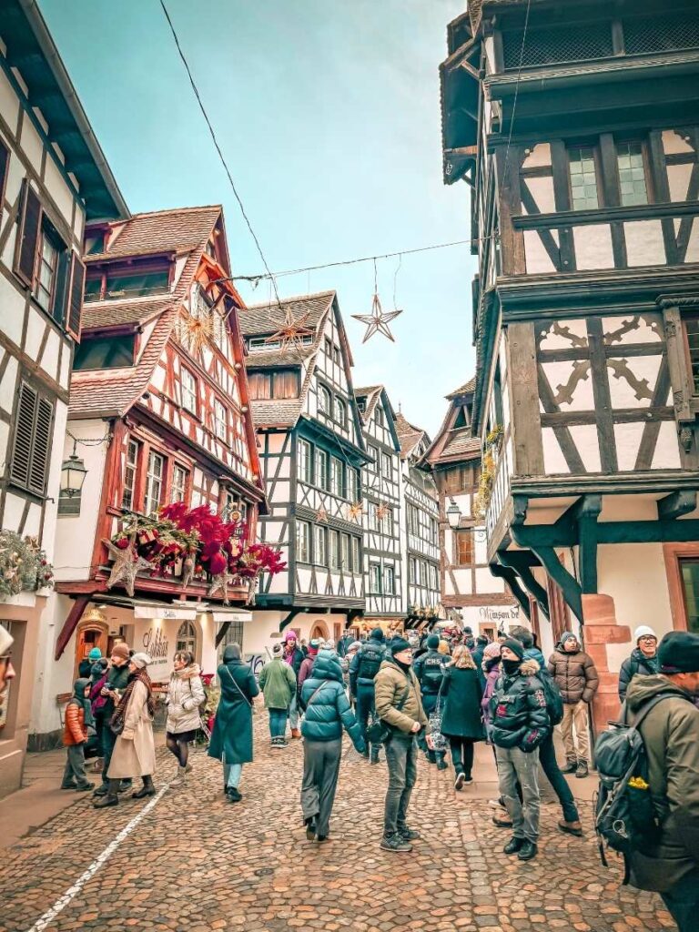 A festive crowd explores the cobbled streets of Petite France, surrounded by half-timbered buildings decorated with red ornaments and stars.