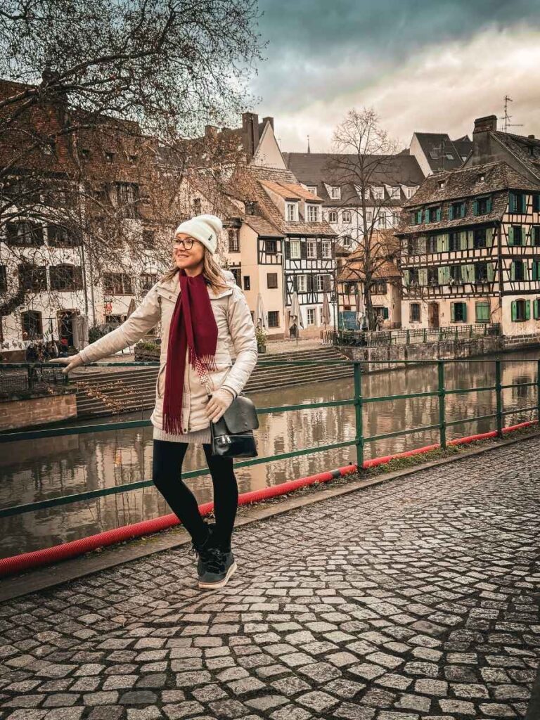 Kate smiles while playfully leaning on a railing beside the canal in Strasbourg’s Petite France, with timbered houses behind her.