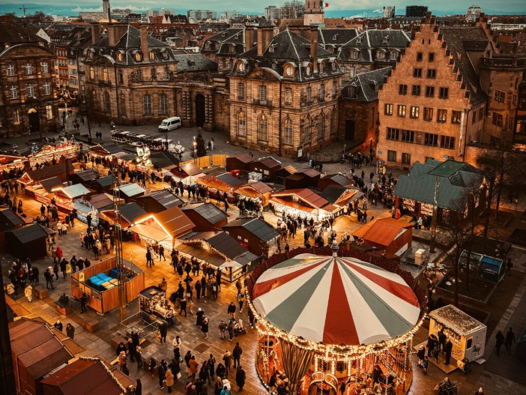 An aerial view of Strasbourg’s Christmas market shows glowing wooden stalls and a striped carousel surrounded by crowds in a historic city square.