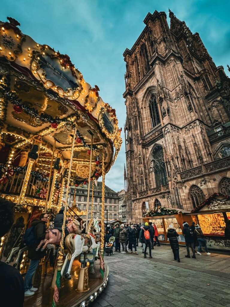 A glowing carousel spins with children riding horses near Strasbourg Cathedral, as marketgoers browse festive stalls beneath the towering Gothic structure.