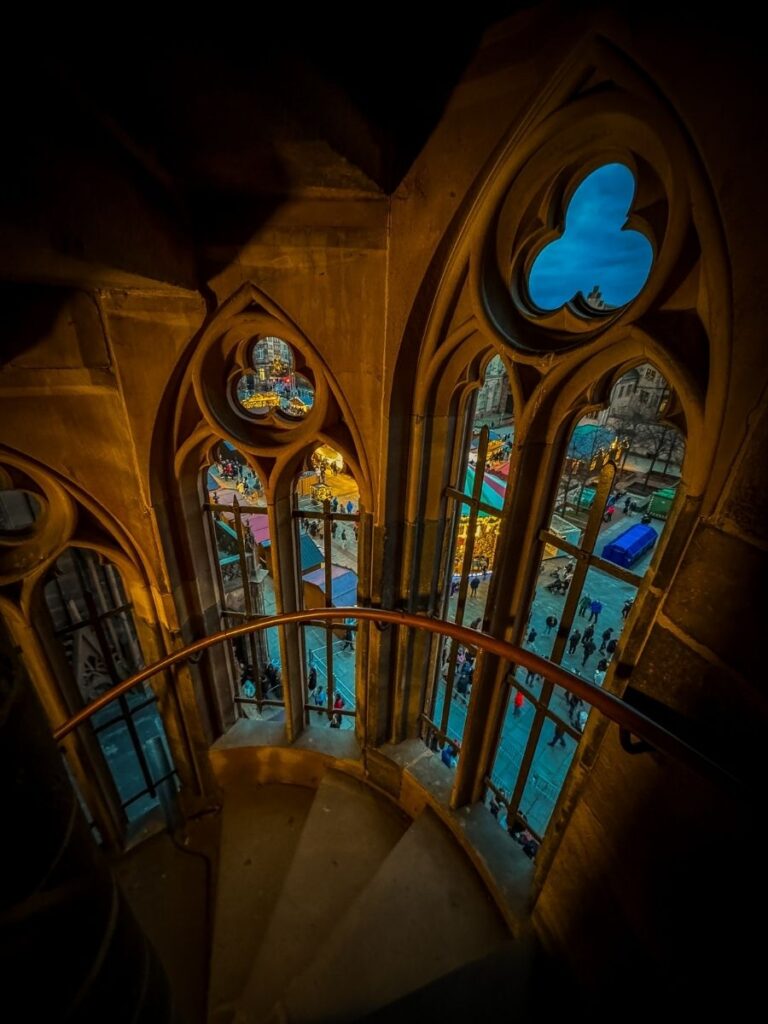 A spiral staircase inside Strasbourg Cathedral frames a view of the glowing Christmas market below through gothic arched windows.