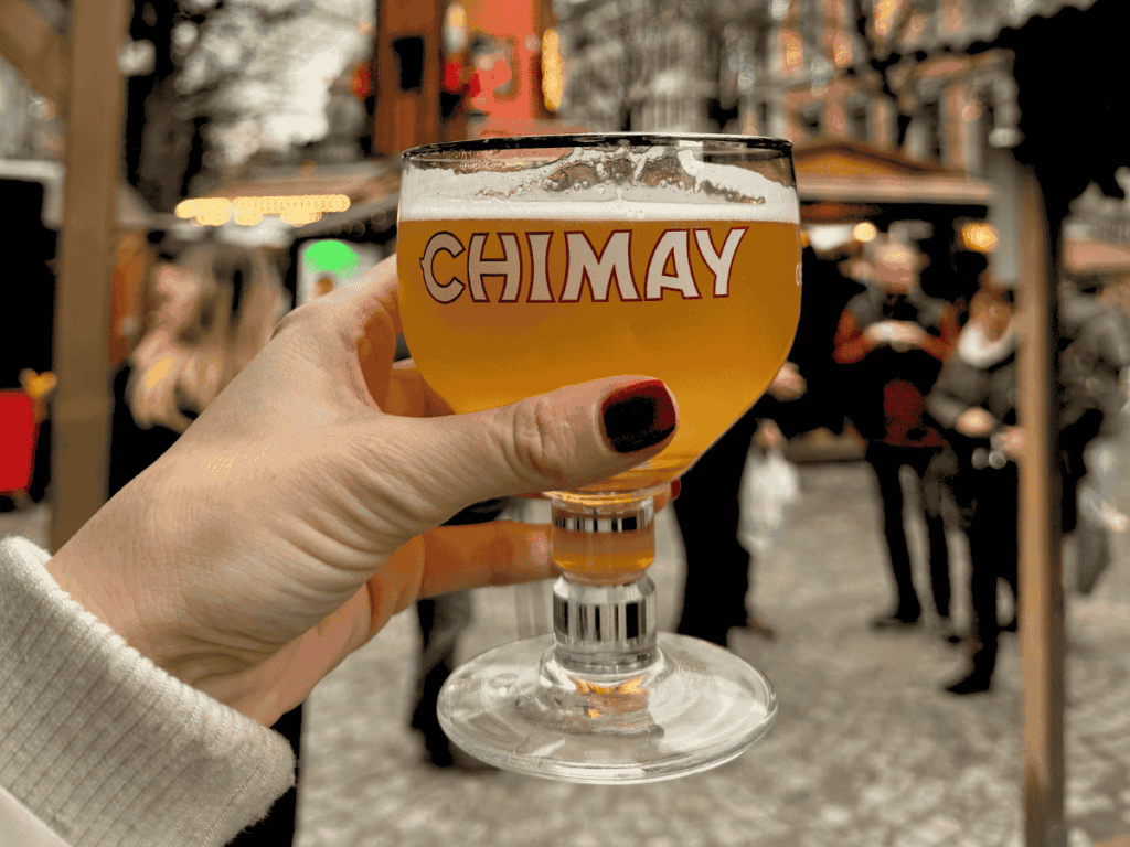 Kate's hand holding up a clear glass of Chimay Belgian Beer.