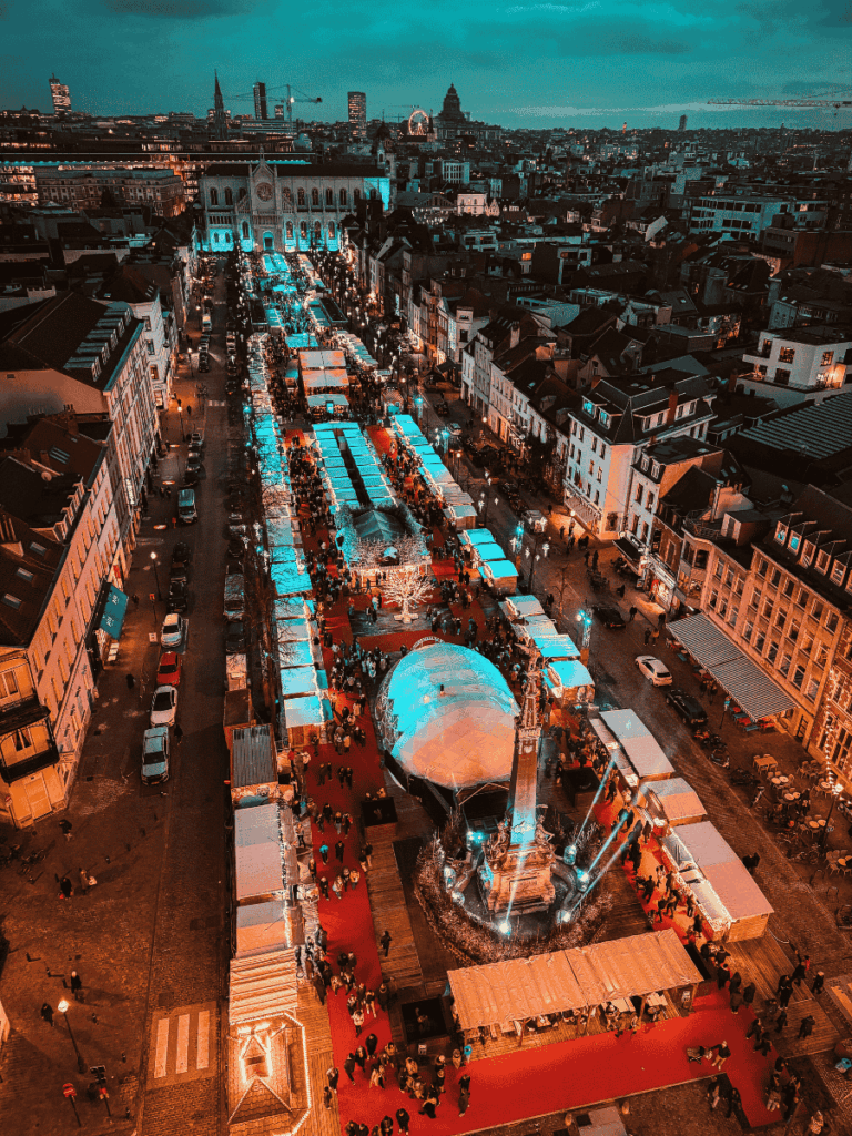 Aerial view of Brussels’ Christmas market glowing with festive lights, red carpets, and busy stalls at dusk.