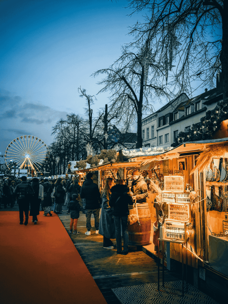 Shoppers browse wooden stalls at Brussels’ Christmas market with a Ferris wheel glowing in the background.