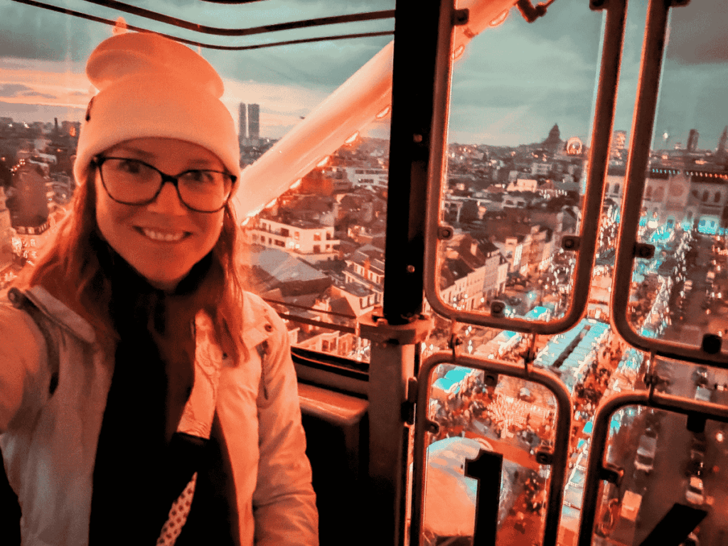 Kate smiles inside a Ferris wheel cabin overlooking the glowing Brussels Christmas market at dusk.