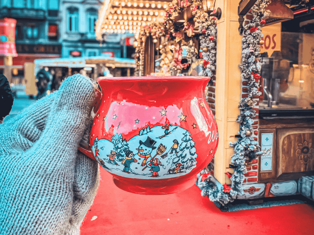 A festive red mug decorated with snowmen and Christmas scenes is held up at a Brussels market stall.