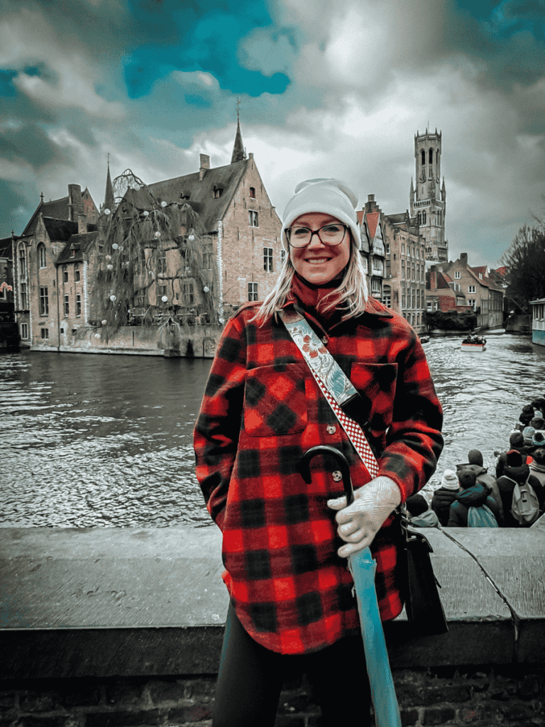 Kate stands by a canal in Bruges with medieval buildings and a tower in the background.