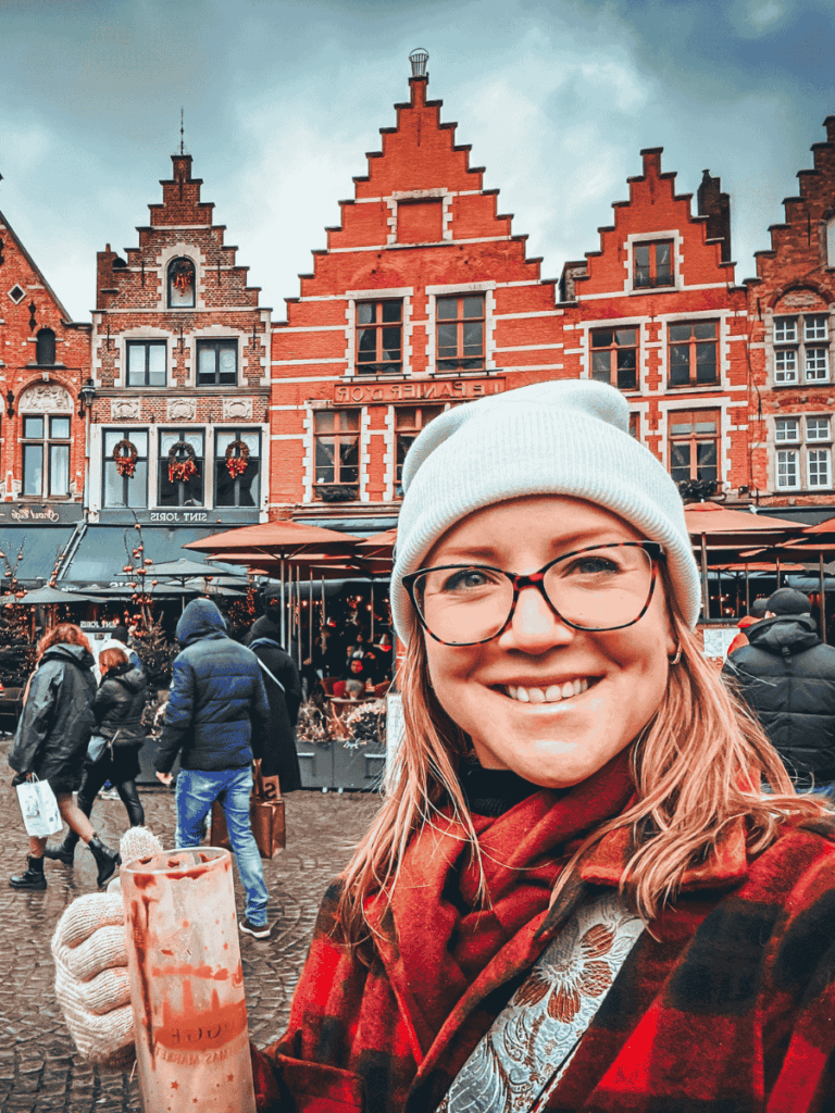 Kate smiles while holding a drink in front of the colorful stepped gable houses of Bruges’ market square.