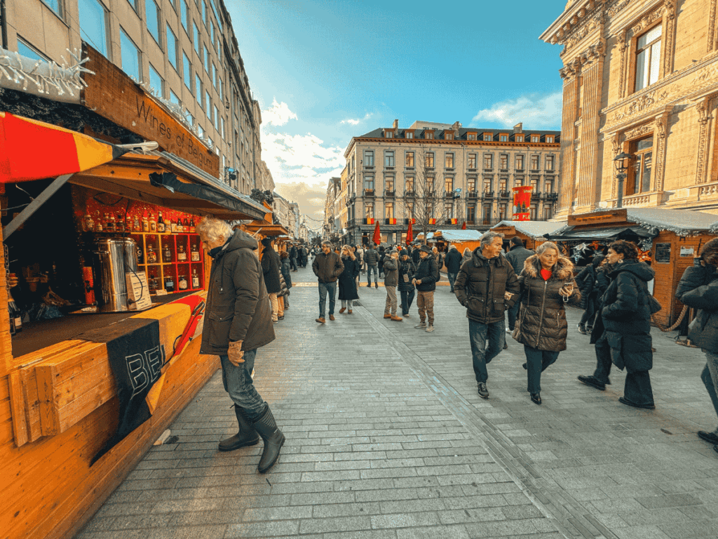 People stroll past wooden stalls at a Christmas market in Brussels on a clear winter day.