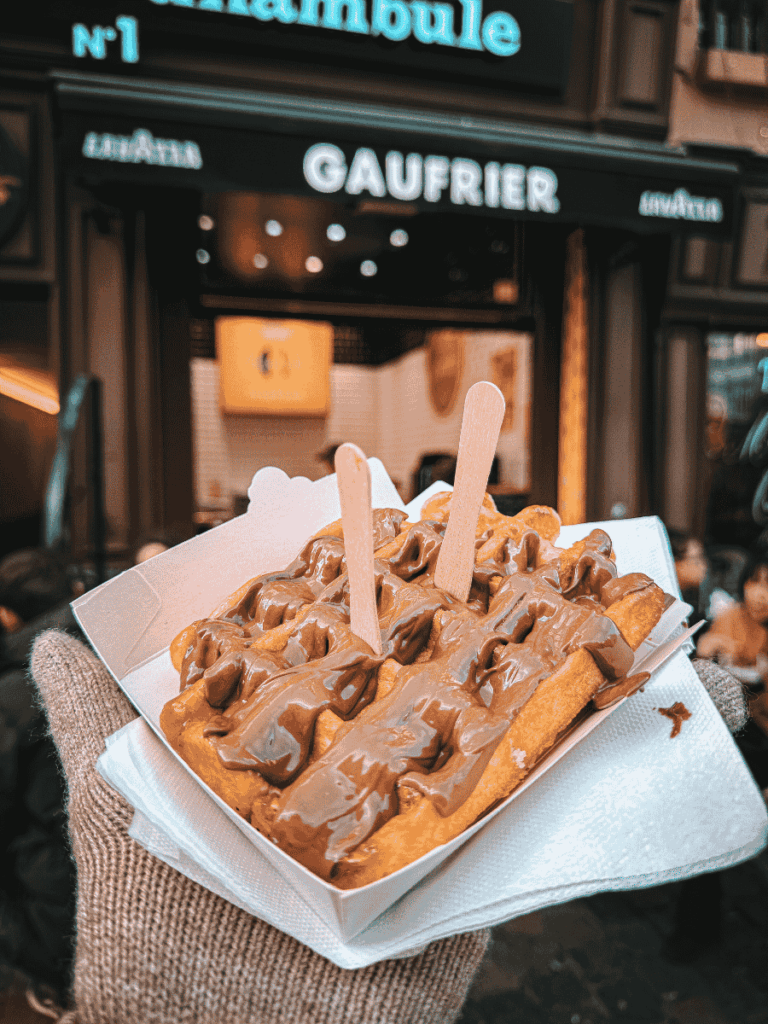 A warm Belgian waffle topped with melted chocolate is held up outside a waffle shop.