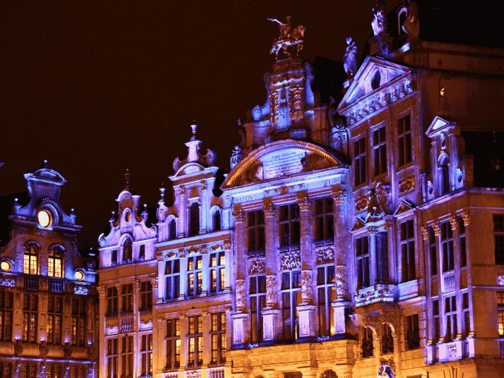Kate smiles under an umbrella in front of a hotel covered in festive Christmas decorations in Bruges.

You said:
She Knows Alt Text said:

Ornate buildings in Brussels’ Grand Place are illuminated with colorful lights at night.