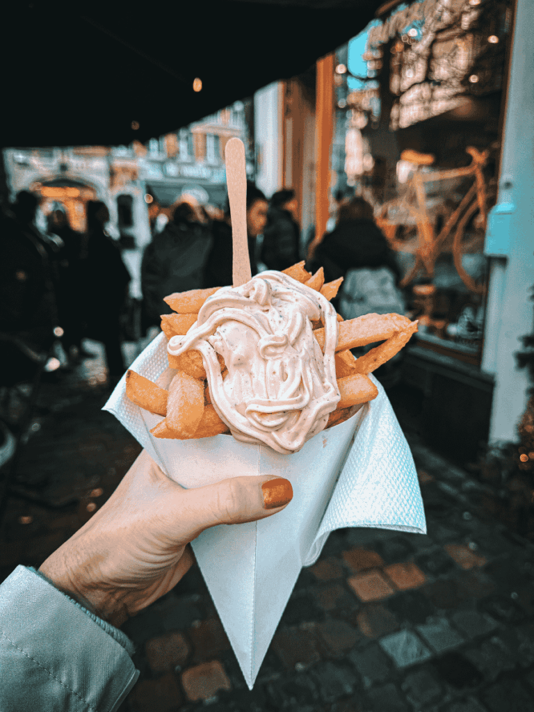 A cone of Belgian fries is topped with a generous swirl of sauce on a busy street.