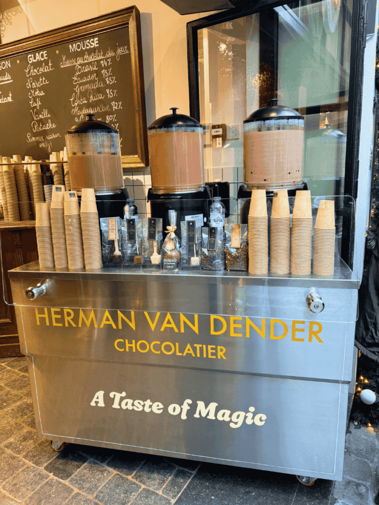 A chocolatier’s counter displays dispensers of rich hot chocolate with stacks of cups ready to serve.